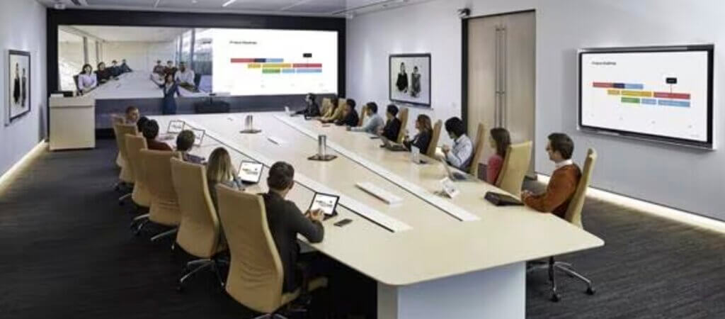 Rendering of a Cisco Redesigned Conference Room. A trapezoid conference table holds attendees in chairs arranged along the two long edges. A screen and camera face one of the shorter edges.